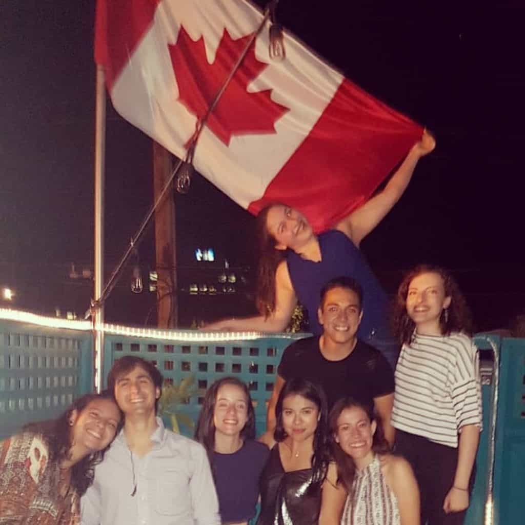 Leticia Teodoro com outros bolsistas do programa Mitacs durante o Canadá Day. Foto com diversos jovens sorridentes à noite segurando uma bandeira do Canadá