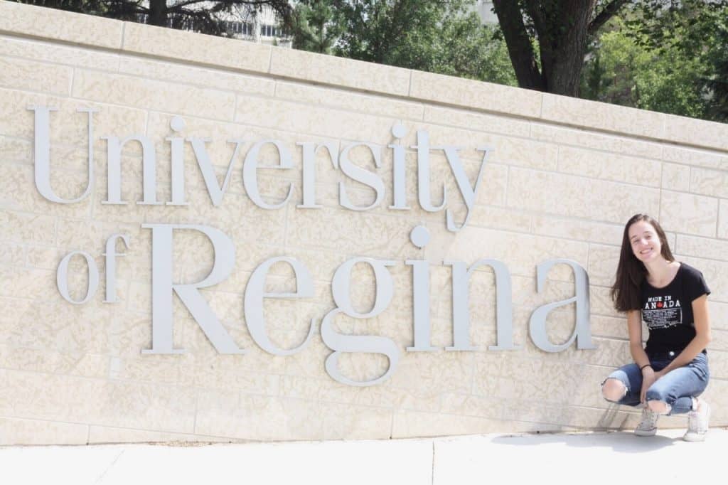 Letícia Teodoro sorri no canto esquerdo na frente do letreiro "university of regina", no Canadá durante o programa Mitacs de estágio para estudantes 