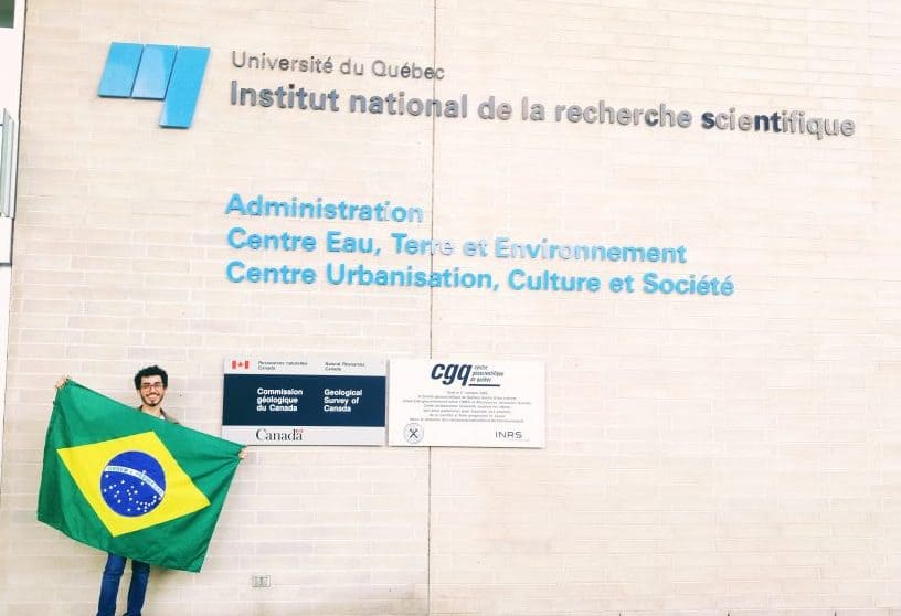 Pedro Ferreira, bolsista do Mitacs Globalink, segura uma bandeira do Brasil na frente da universidade do Quebec e dá dicas de como fazer estágio em biotecnologia no Canadá