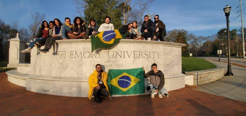 FLTAs Brasileiros sentados em um letreiro da Emory University nos EUA segurando uma bandeira dos Brasil