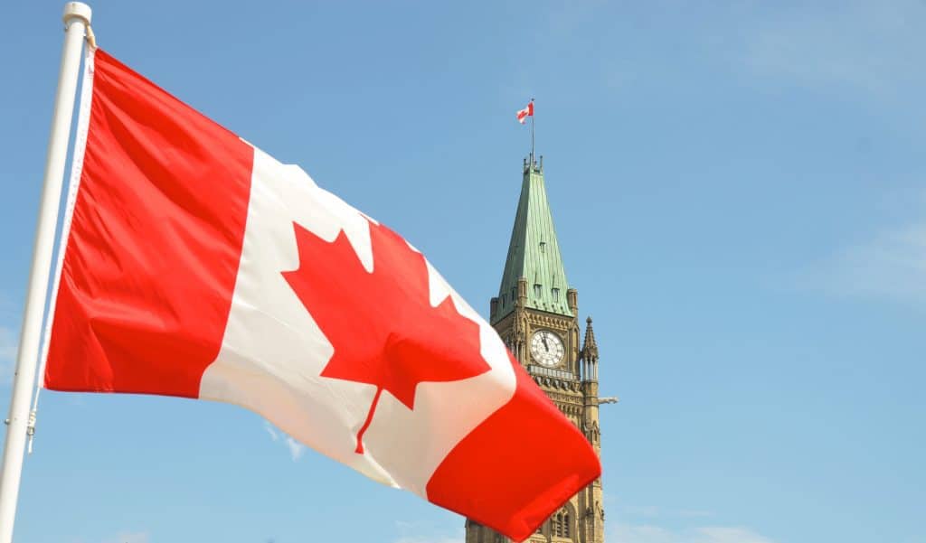 Foto mostra a bandeira do canadá ao canto esquerdo e um prédio antigo com relógio no cando direito e céu azul bolsa de estudos Canadá partiu intercambio