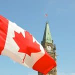 Foto mostra a bandeira do canadá ao canto esquerdo e um prédio antigo com relógio no cando direito e céu azul bolsa de estudos Canadá partiu intercambio